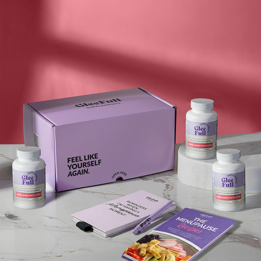 Subscription bundle display for GleeFull Meno-Shred on a marble surface against a pink background. The layout includes a large purple shipping box reading 'FEEL LIKE YOURSELF AGAIN.' and 'OPEN HERE', three white bottles of Meno-Shred Fat Burner, a purple branded pen, a matching notepad that reads 'REMINDERS WHEN I'M HAVING A MENOPAUSE MOMENT', and a recipe booklet titled 'THE MENOPAUSE Recipes'.