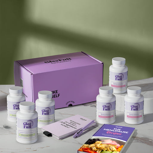 A product bundle display for GleeFull Supplements on a marble surface against a green background. The layout includes a large purple shipping box featuring the slogan 'FEEL LIKE YOURSELF AGAIN.', three white bottles of Over 30 Hormone Support and three white bottles of Meno Joint Relief, a purple branded pen, a matching notepad that reads 'REMINDERS WHEN I'M HAVING A MENOPAUSE MOMENT', and a recipe booklet titled 'THE MENOPAUSE Recipes'