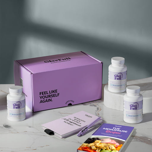 A product bundle display for GleeFull Supplements on a marble surface against a blue background. The layout includes a large purple shipping box featuring the slogan 'FEEL LIKE YOURSELF AGAIN.', three white bottles of Bloat Buster, a purple branded pen, a matching notepad that reads 'REMINDERS WHEN I'M HAVING A MENOPAUSE MOMENT', and a recipe booklet titled 'THE MENOPAUSE Recipes'