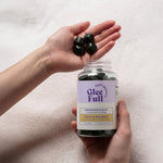 
A person holds a clear GleeFull Calm & Balance bottle with dark gummies in their open palm, against a soft cream blanket. Label reads 'Ashwagandha Root, Calm & Balance, 60 Gummies'.