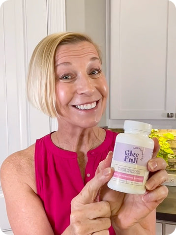 A smiling blonde woman in a bright pink top holds up a white bottle of Glee Full dietary supplement in front of a white kitchen background.